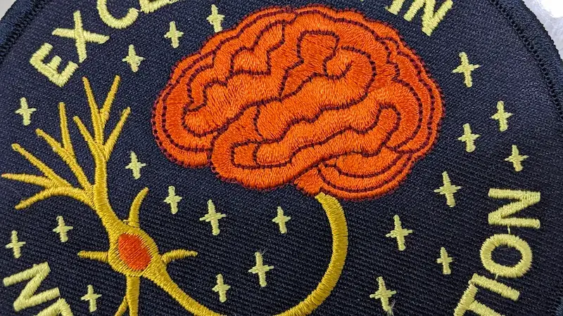 Close up of a custom embroidered brain patch showing intricate stitching, fine text, and high-detail thread work.